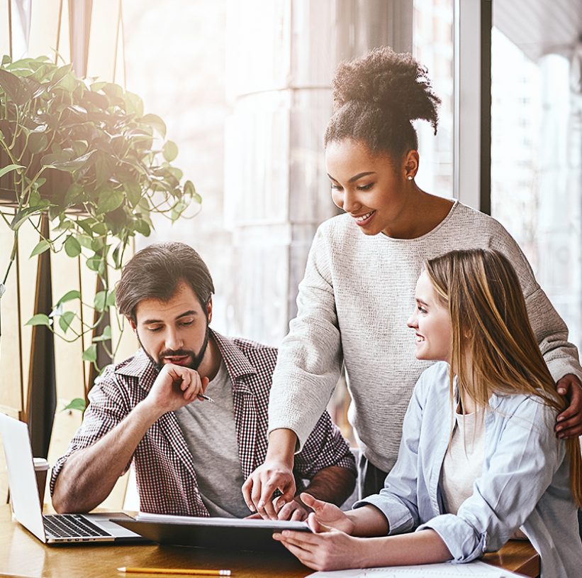 Three cheerful colleagues discussing new ideas in office. Bearded man looks on the chart, while women talk to him. A laptop computer, notebook and pencils are on the desk. Casual style. Concept of success